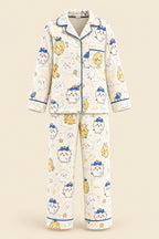 A woman takes a mirror selfie wearing a two-piece cartoon pajama set with a cream base and blue, yellow, and pink printed characters. She is sitting on a beige couch with plush toys in the background.