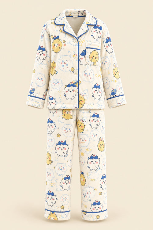A woman takes a mirror selfie wearing a two-piece cartoon pajama set with a cream base and blue, yellow, and pink printed characters. She is sitting on a beige couch with plush toys in the background.